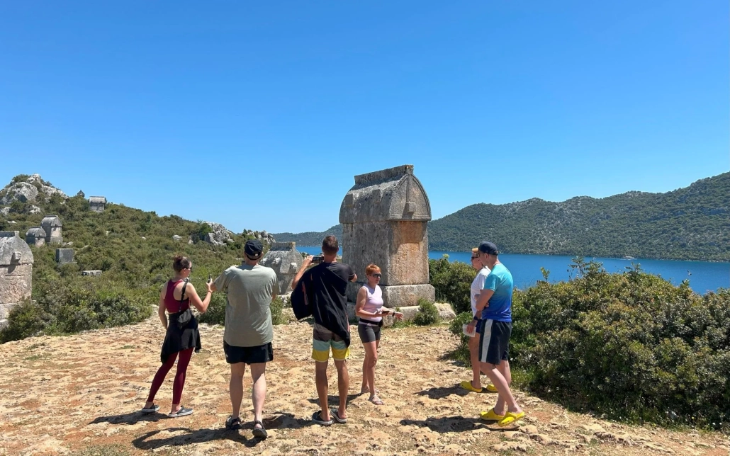 A group of travelers exploring ancient Lycian rock tombs on a hilltop above the sea in Kekova, with panoramic views of the bay in the background.