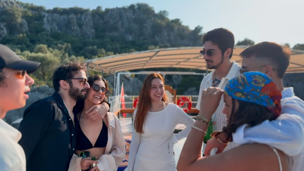 A group of young friends smiling and enjoying themselves on the deck of a boat during a Kekova tour, with forested hills in the background.