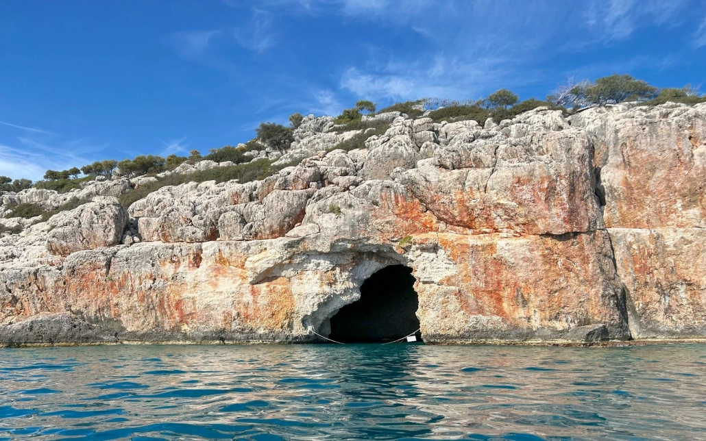 Entrance of Pirate Cave along the rocky sea cliffs of Kekova, Turkey, with turquoise water and a bright sky.