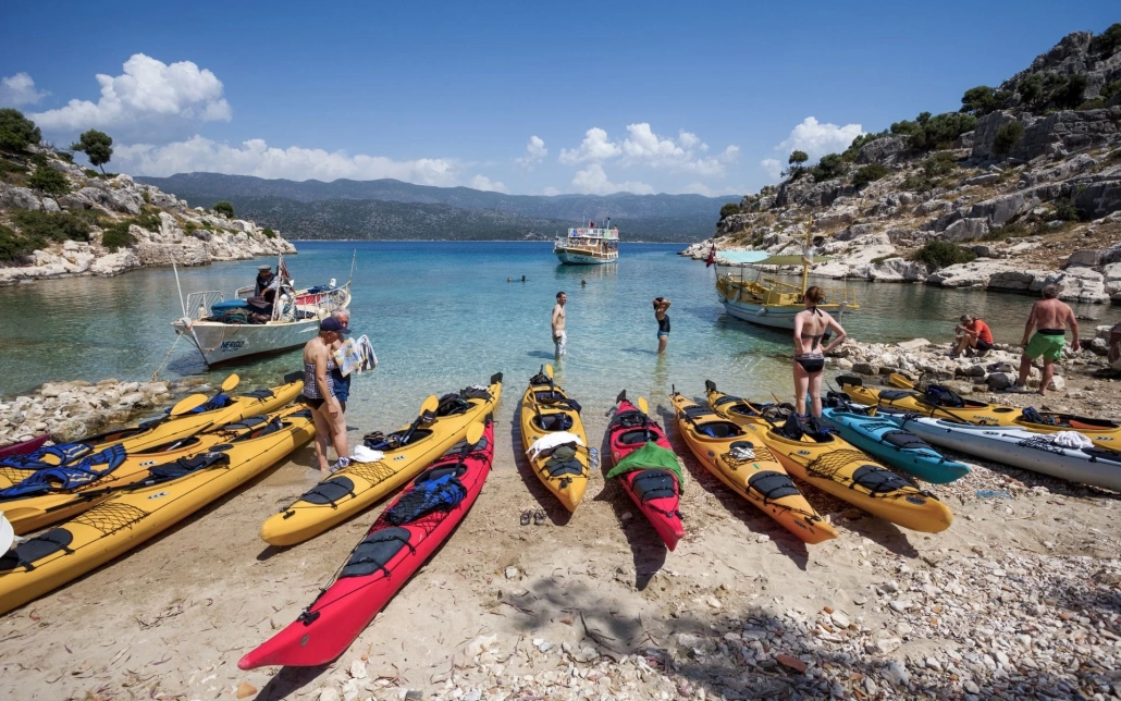 A group of people taking a break with colorful sea kayaks lined up on the beach in Kekova, Antalya, with boats anchored in the turquoise bay.