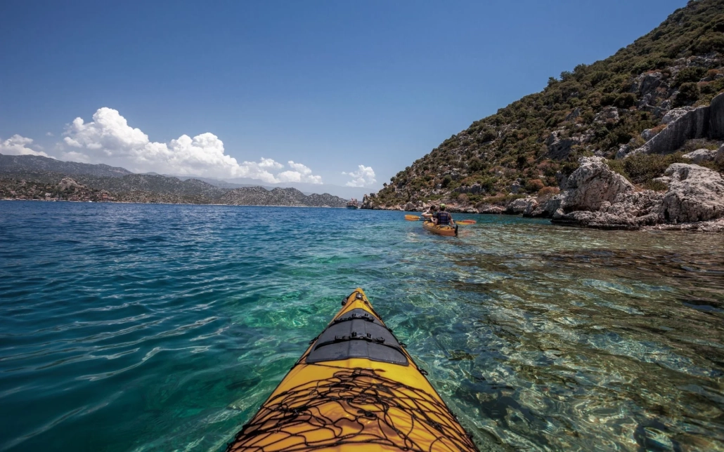 Paddling a yellow sea kayak along the scenic coastline of Kekova, Antalya, with clear turquoise water and rocky hills in the background.