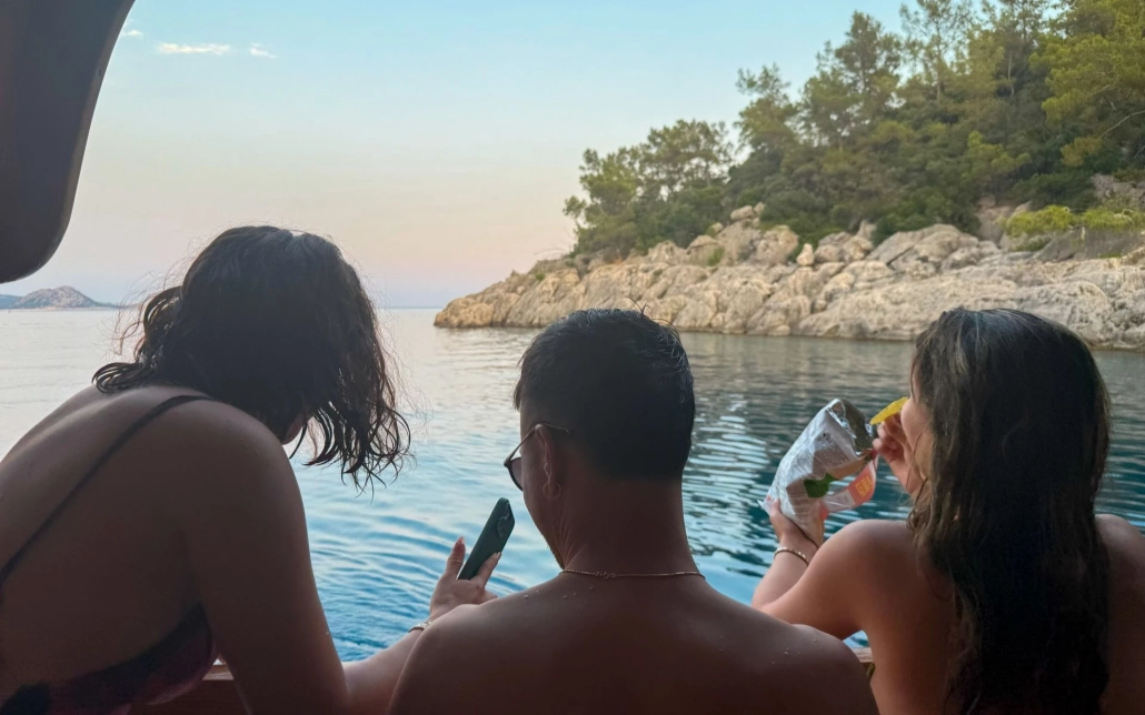Three young adults enjoying snacks and relaxing on the back of a boat during a Blue Cruise in Kekova, with rocky coastline and calm water at sunset.